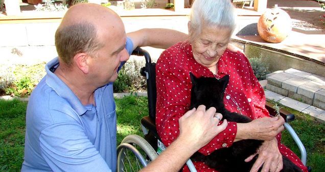 Seniorin im Rollstuhl hält Katze, mit knieendem Pfleger an ihrer Seite im Garten