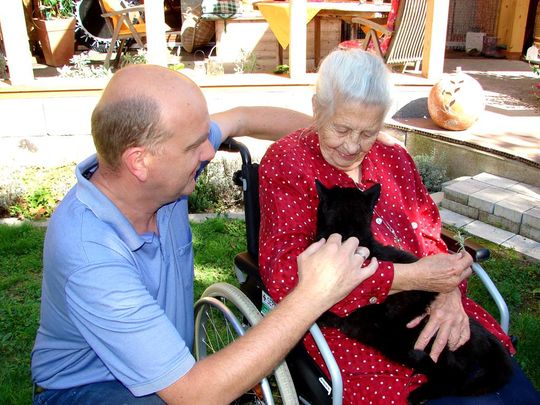 Seniorin im Rollstuhl hält Katze, mit knieendem Pfleger an ihrer Seite im Garten